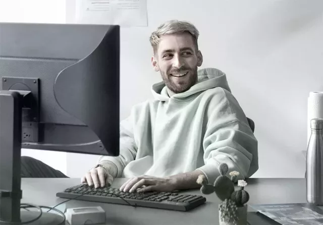 Ein Agentur-Mitarbeiter in einem hellgrünen Kapuzenpulli sitzt lächelnd an einem Schreibtisch, beide Hände auf der Tastatur, vor einem Computermonitor.
