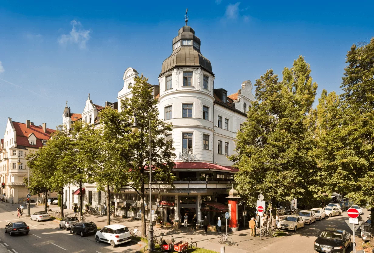 Das Feinkost Käfer Stammhaus in der Prinzregentenstraße in München. Ein historisches Gebäude an einer Straßenecke mit in der Nähe geparkten Autos, umgeben von Bäumen unter einem blauen Himmel.