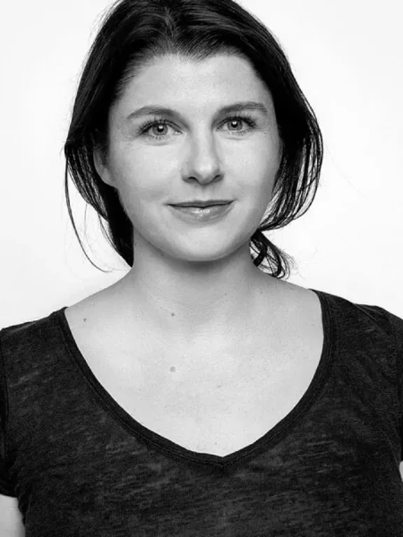 Frau mit dunklem Haar, trägt ein schwarzes T-Shirt und blickt vor einem schlichten Hintergrund direkt in die Kamera.