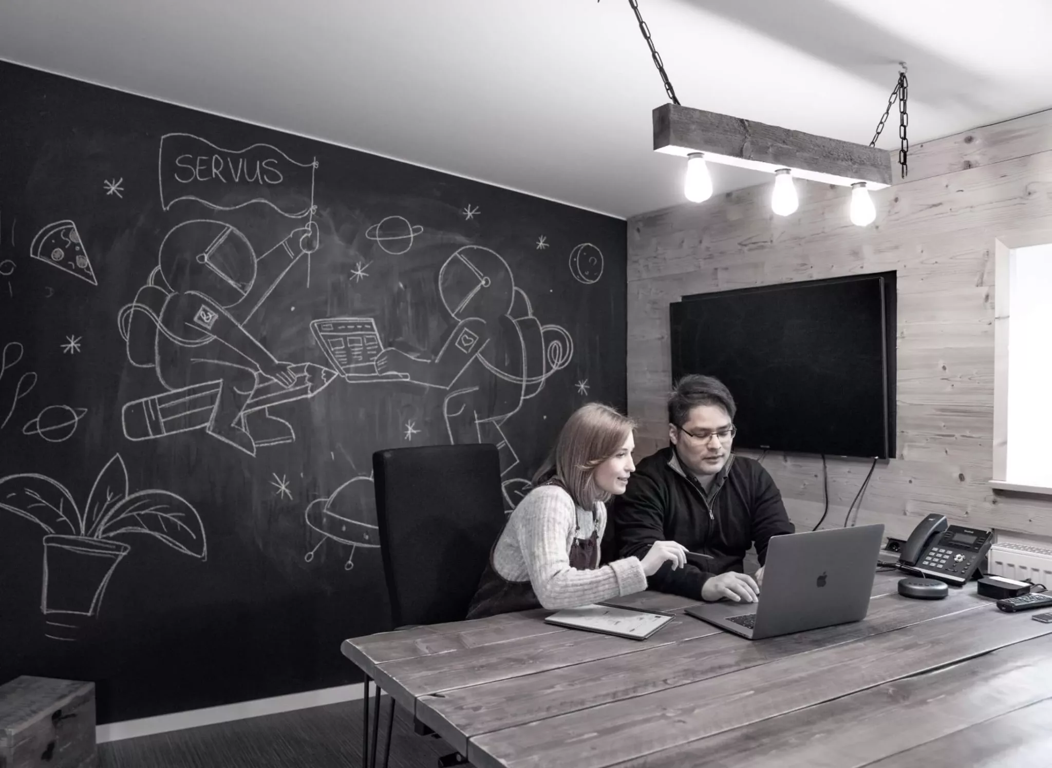 Eine Frau und ein Mann sitzen vertieft zusammen am Laptop in einem Konferenzraum, der an eine Blockhütte erinnert, und diskutieren ein Website Konzept. Im Hintergrund sind an eine Kreidetafel zwei Astronauten gezeichnet. Einer der beiden schwenkt eine Flagge, auf der "Servus" geschrieben steht.