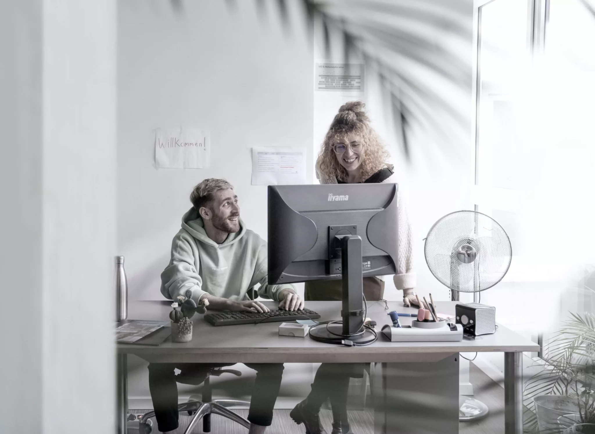 Zwei Personen arbeiten gemeinsam an einem Schreibtisch mit einem Computer in einem hellen Büro. Eine Person sitzt und tippt, während die andere steht und sich vorbeugt. Beide schauen auf den Bildschirm und lächeln.
