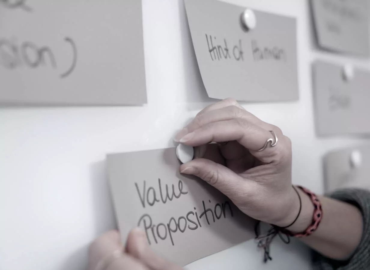 Die Hand einer Frau pinnt mit einem Magnet ein Kärtchen auf dem "Value Proposition" steht an ein Whiteboard. Im Hintergrund sind verschwommen weitere Kärtchen zu sehen.