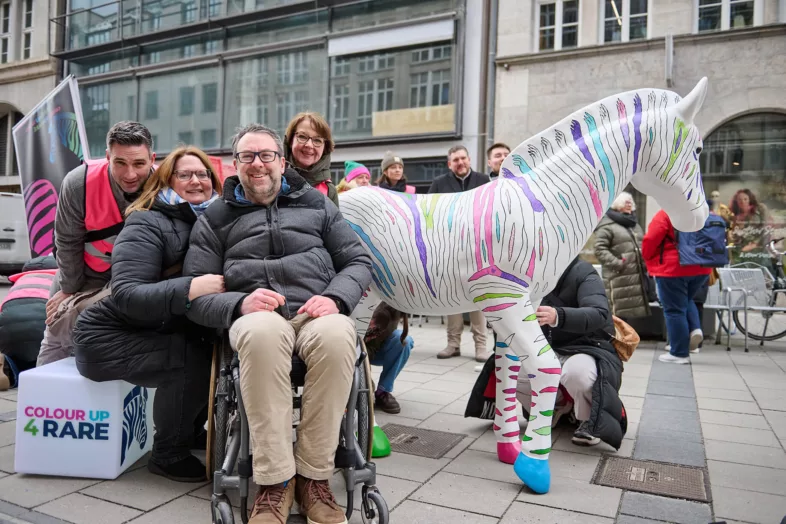 Ein Mann im Rollstuhl, der sich mit seiner Familie vor der bunten Zebra-Skulptur fotografieren lässt.