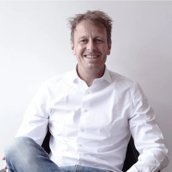Matthias Bierich, Geschäftsführer bei schalk&friends, der in einem weißen Hemd und Jeans locker auf einem Stuhl sitzt und im die Kamera lächelt.