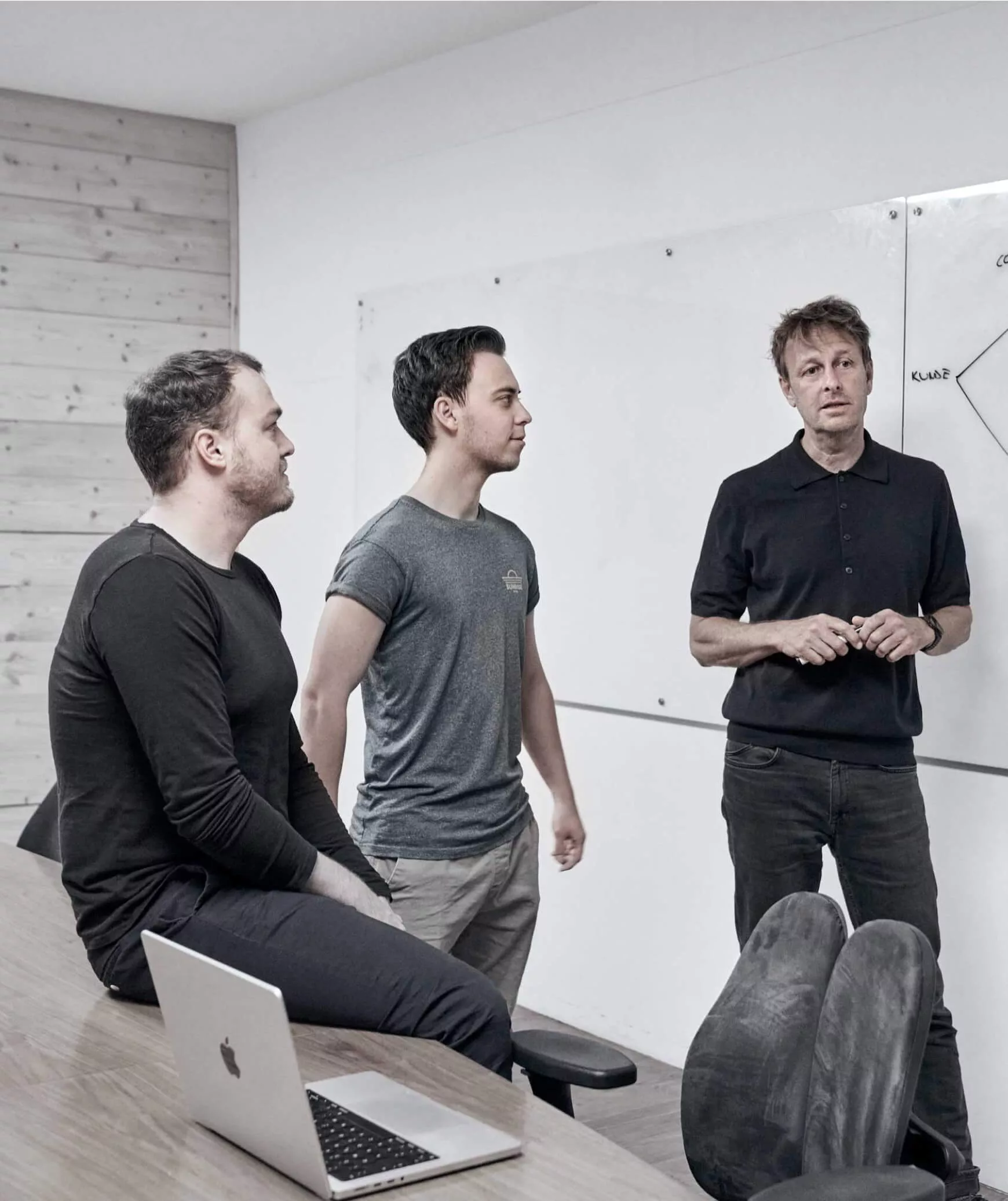 Drei Männer führen in einem Besprechungsraum einen KI Experience Workshop durch; einer steht an einer Tafel mit Diagrammen, während die beiden anderen an einem Tisch mit einem Laptop sitzen und zuhören.