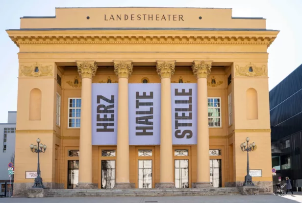 Die Fassade eines gelben klassizistischen Gebäudes mit der Aufschrift "Landestheater", zwischen dessen Säulen drei große Banner mit der Aufschrift "HERZ HALTE STELLE" hängen.