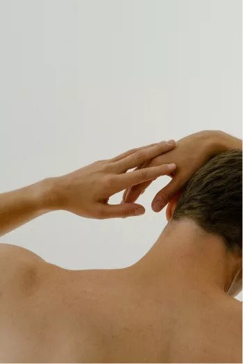 Eine Person mit kurzen braunen Haaren wird von hinten gezeigt, die ihren linken Arm hebt und ihren Hinterkopf vor einem schlichten, hellen Hintergrund berührt.