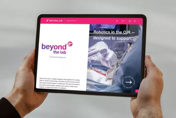 Eine Person, die ein Tablet in der Hand hält, auf dem das digitale Magazin mit der Überschrift „Robotik im OP – zur Unterstützung konzipiert“ und dem Logo „beyond the lab, The Brainlab Magazine.“ zu sehen ist.